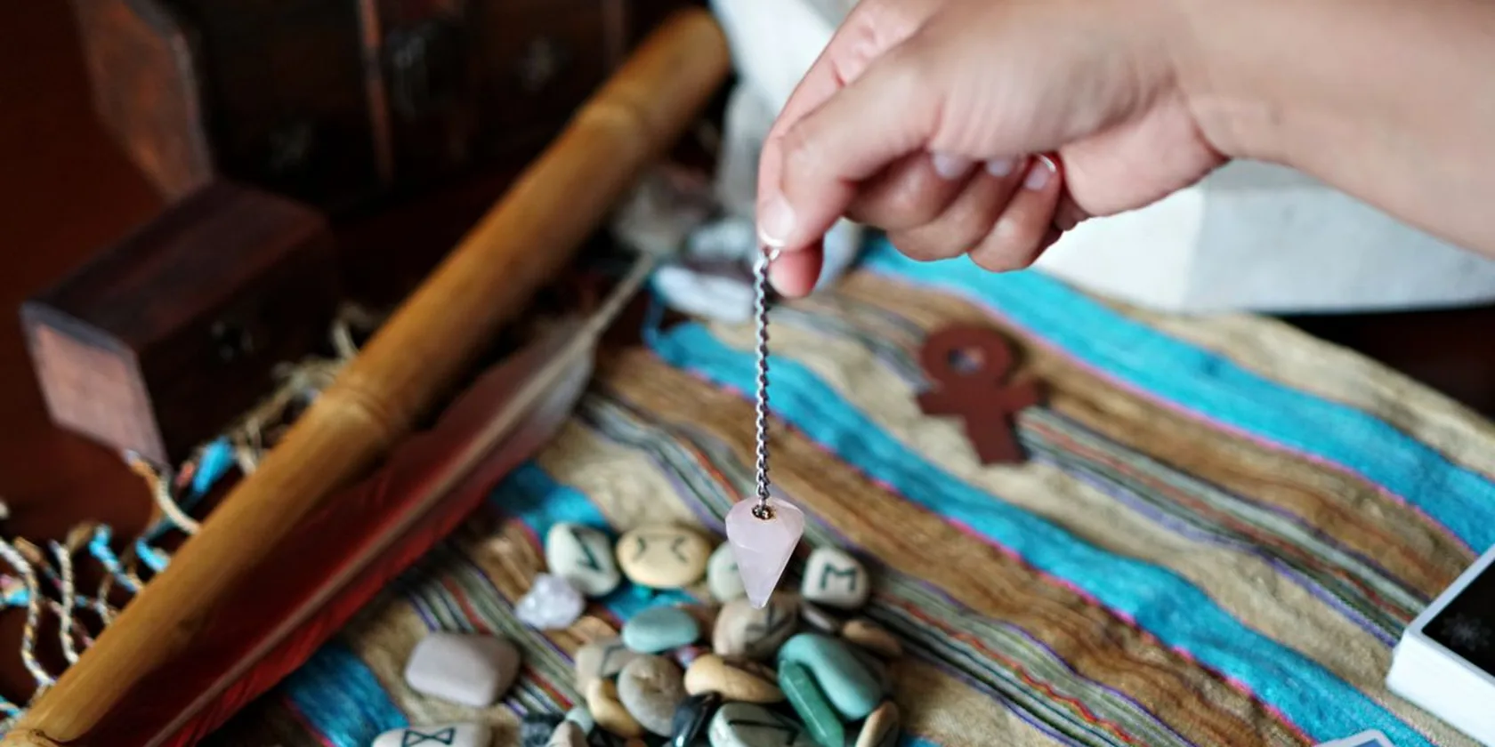 Pendule de radiesthésie en quartz rose suspendu au-dessus de runes et cartes de tarot
